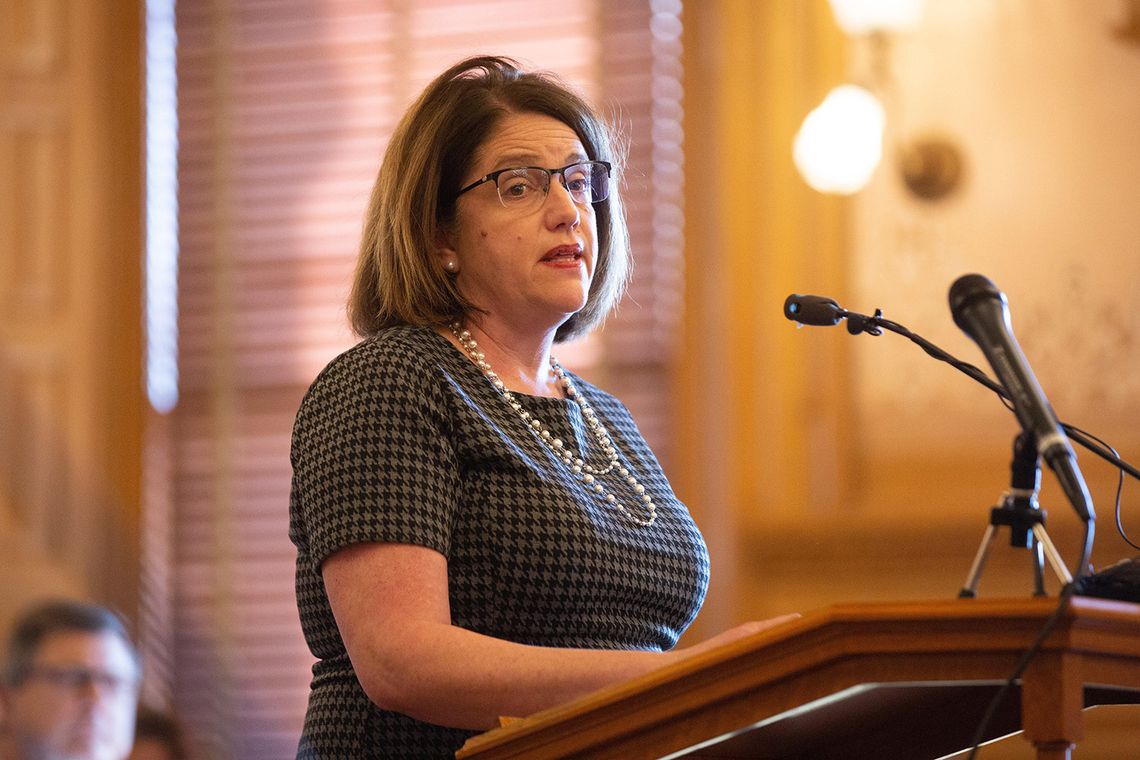 A White woman with medium-length light brown hair wearing glasses, stands at a lectern. 
