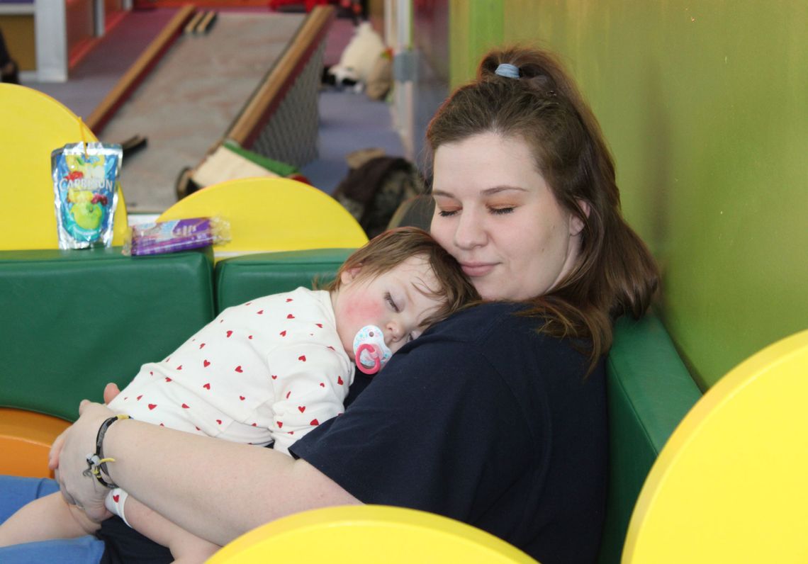 A White woman in a black T-shirt sits on a green couch, holding her baby daughter on her chest, as the little girl sleeps with a pacifier in her mouth. 