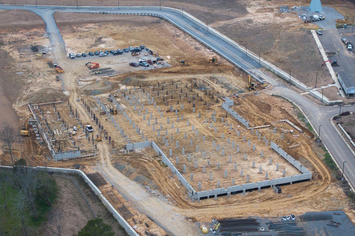 An aerial photo shows a site in the early stages of construction. Cars are parked in a lot near the site. 