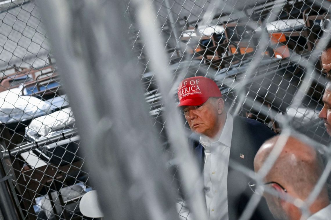 A photograph of a White man in his late 70s wearing a red hat with the words "Gulf of America" and a suit. He is seen through an out-of-focus chainlink fence and is looking to the side. Behind him there are rows of bunk beds behind another chainlink fence. 