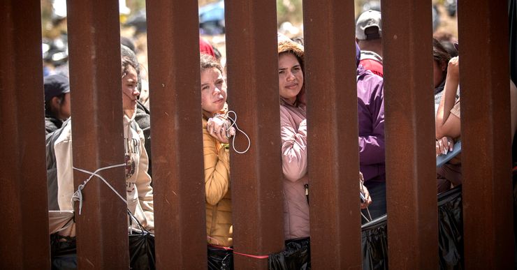 Two women with medium skin tone, holding onto their belongings, stand in between brown pillars of a wall while waiting in a line. Several people stand in the background. 