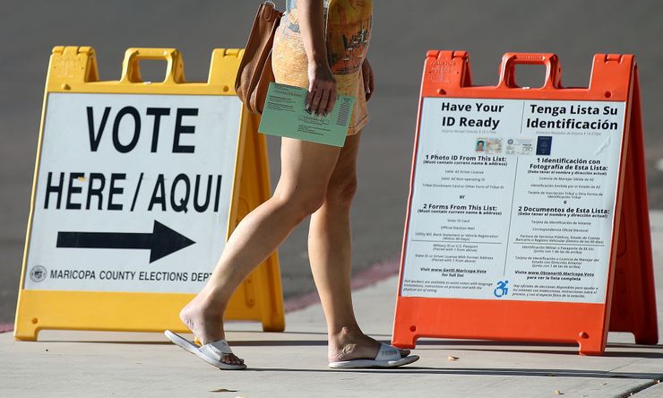 A photo shows a person walking with their ballot. In the background, there are two voting signs. One sign reads, "Vote Here/Aqui, Maricopa County Elections Dept." The second sign provides photo ID details in English and Spanish. 