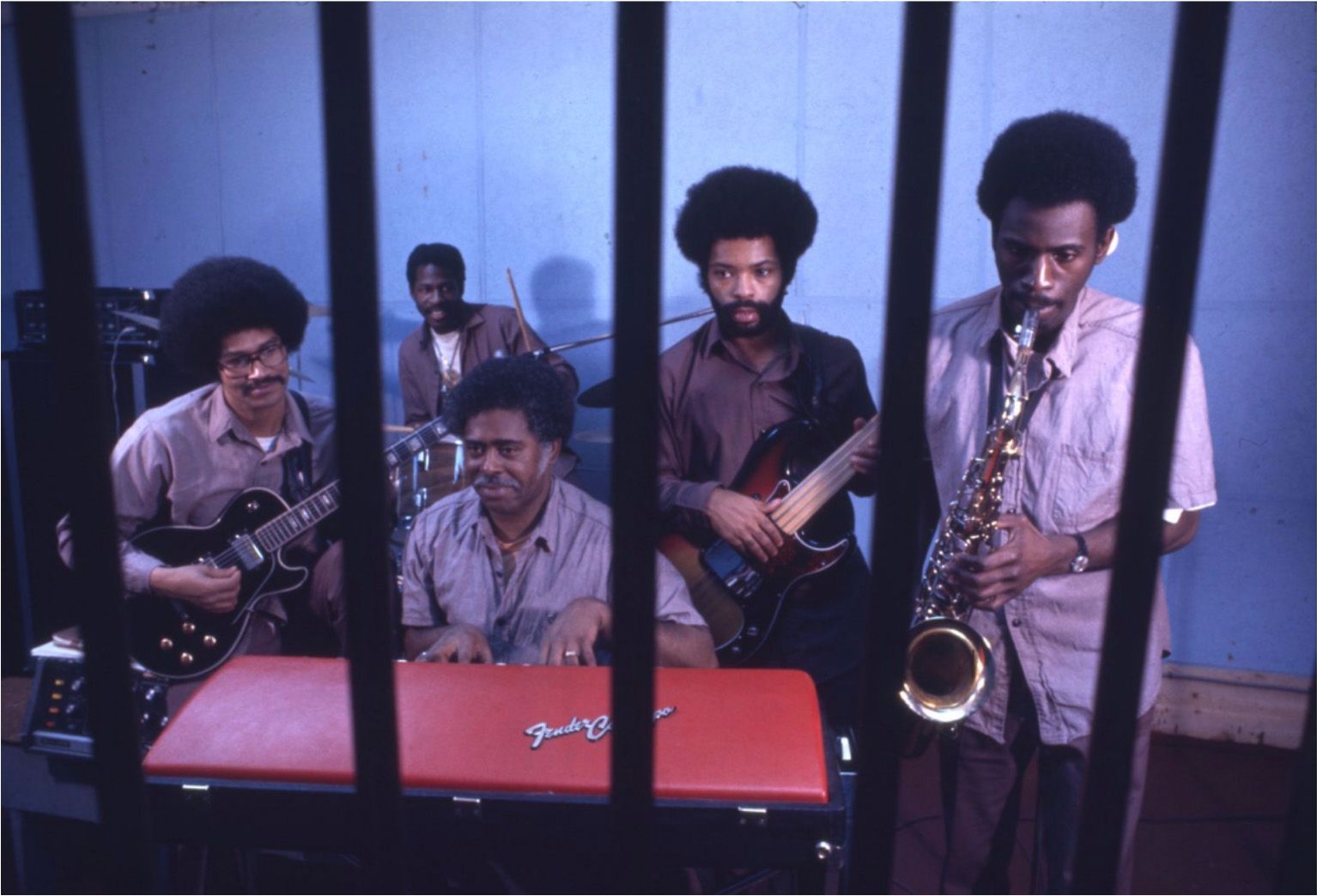 A musical band of Black men are playing instruments, such as a saxophone, guitar, piano, bass guitar and drums. They play behind bars in a room. 