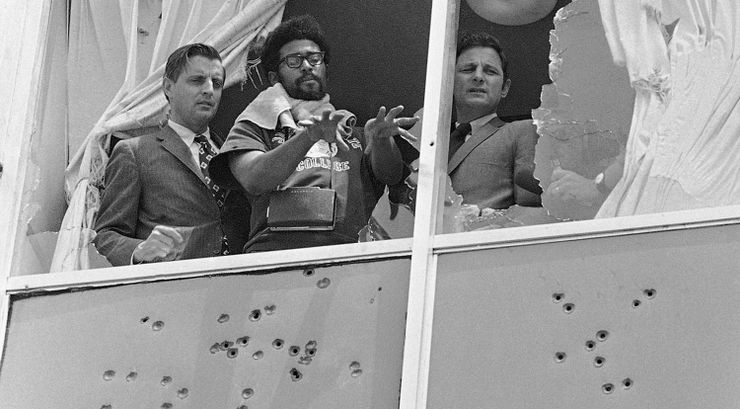 A black-and-white photograph shows two White men in suits looking out a shattered window, while a Black man in a T-shirt gestures downward. The parts of the building around the window are covered in bullet holes. 
