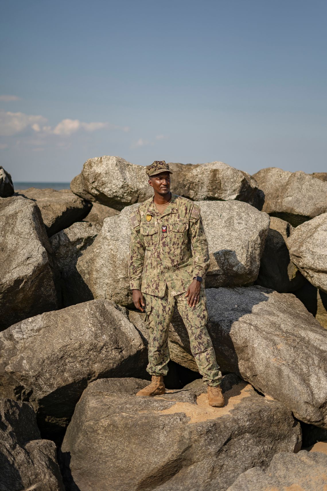 Vincent Montague, a Black man, wears a military uniform while standing on a rock against a wall of rocks with the sea visible behind them. 