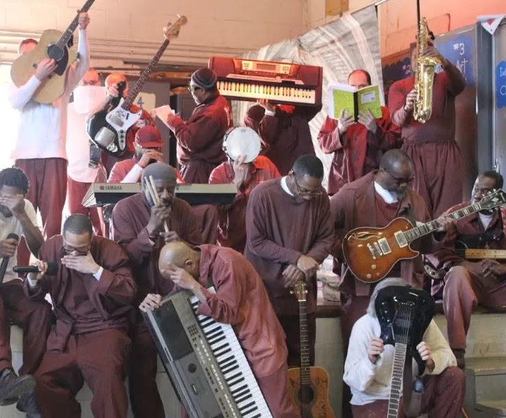 A photo shows a group of over a dozen men, holding their instruments and one case, a microphone. One is holding a keyboard, a few are holding guitars, one is holding a saxophone, and one a book. They are mostly dressed in maroon uniforms. 