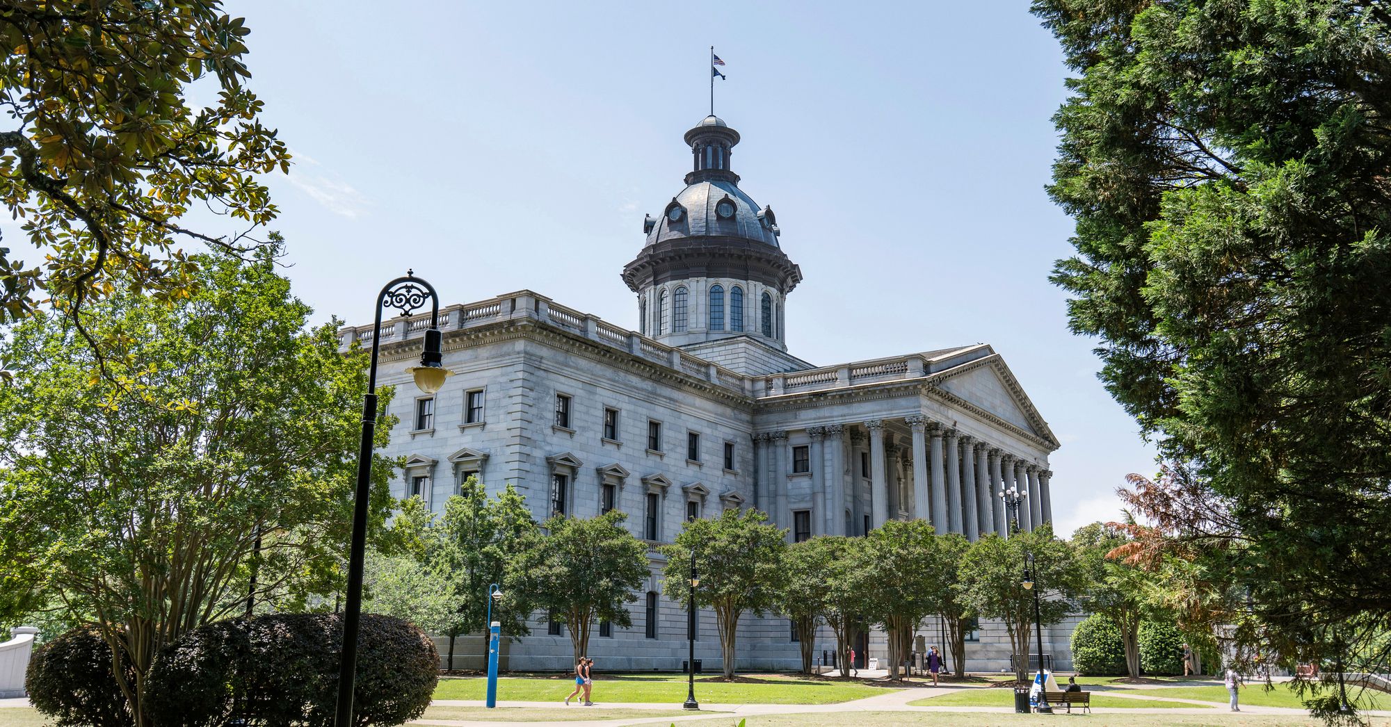 A photograph from an angle of the South Carolina State House, a light-colored building with a triangular pediment and columns in front, and trees on a lawn with paths. 