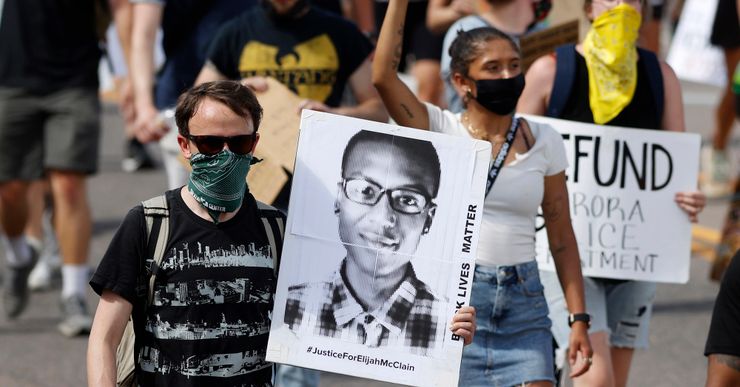 A White person, wearing sunglasses, a green bandana mask and black shirt, holds a poster with the black-and-white image of Elijah McClain, a Black man wearing glasses and a plaid button-up shirt. The poster also reads, "Black Lives Matter. #JusticeForElijahMcClain." There are more protesters marching in the background. 