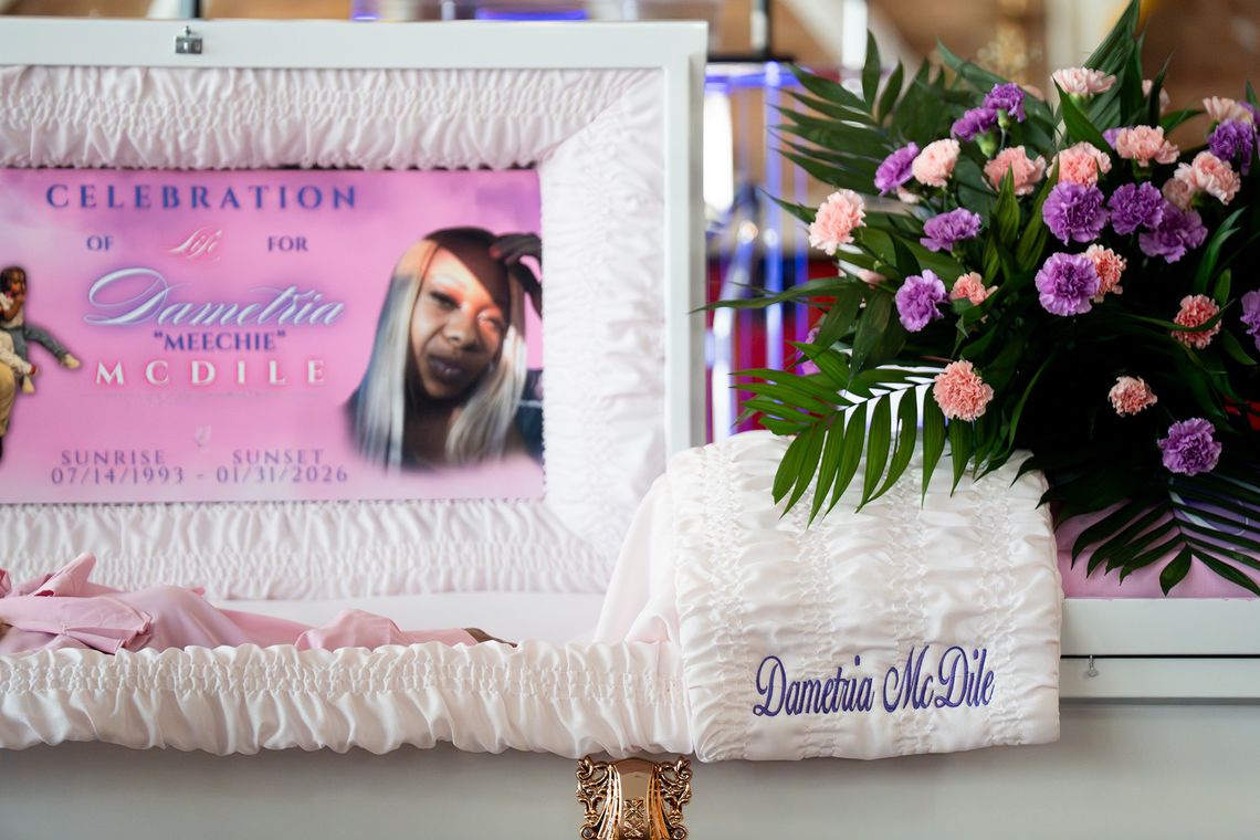 A photo shows an open white casket with the name “Dametria McDile” embroidered on white fabric and a floral arrangement placed on top. The deceased person inside the casket is wearing pink clothing. A pink poster with an image of a Black woman inside the casket reads, “Celebration of Life for Dametria ‘Meechie’” McDile, Sunrise 7/14/1993 - Sunset 1/31/2026.” 
