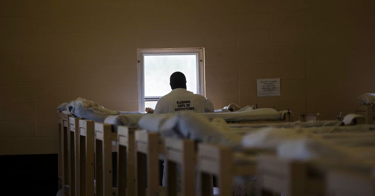 A man in a white prison uniform with the label "Alabama Dept of Corrections" on his back stands near his bunk bed. There are multiple bunk beds lined up in the foreground.