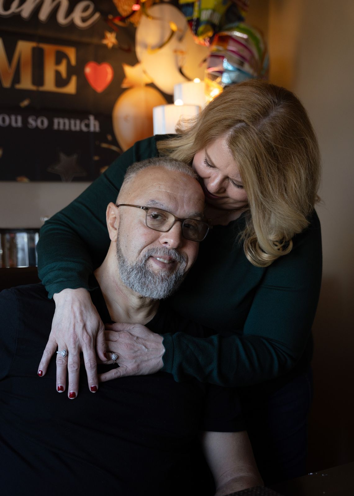 A photo shows Sandra, a White woman, hugging Latif Hafraoui, a man with medium-toned skin. Latif has a gray beard and glasses and is wearing a black t-shirt. Sandra is wearing a green long-sleeved shirt and has medium-length blonde hair. 