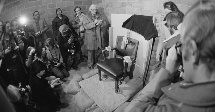 A black and white photo shows a black chair draped with a hood which Gilmore wore during the execution. The upper right part of the chair back has bullet holes. The chair is surrounded with sandbags. 