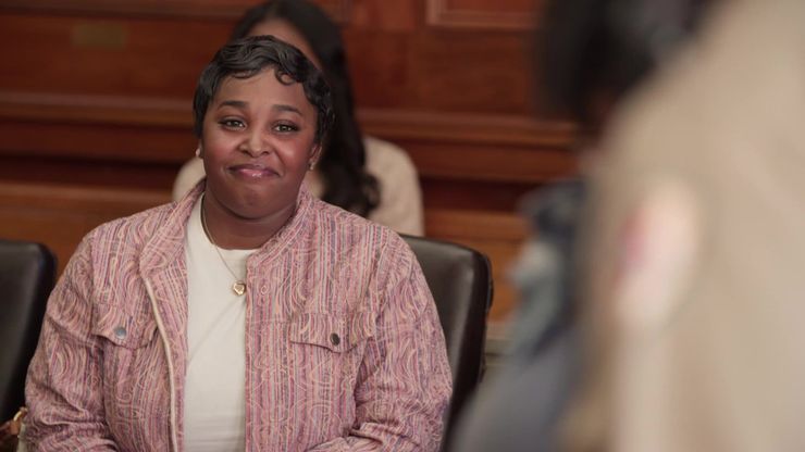 A Black woman with short hair,  wearing a patterned-pink jacket, sits in a chair in a courtroom with a small smile on her face. 