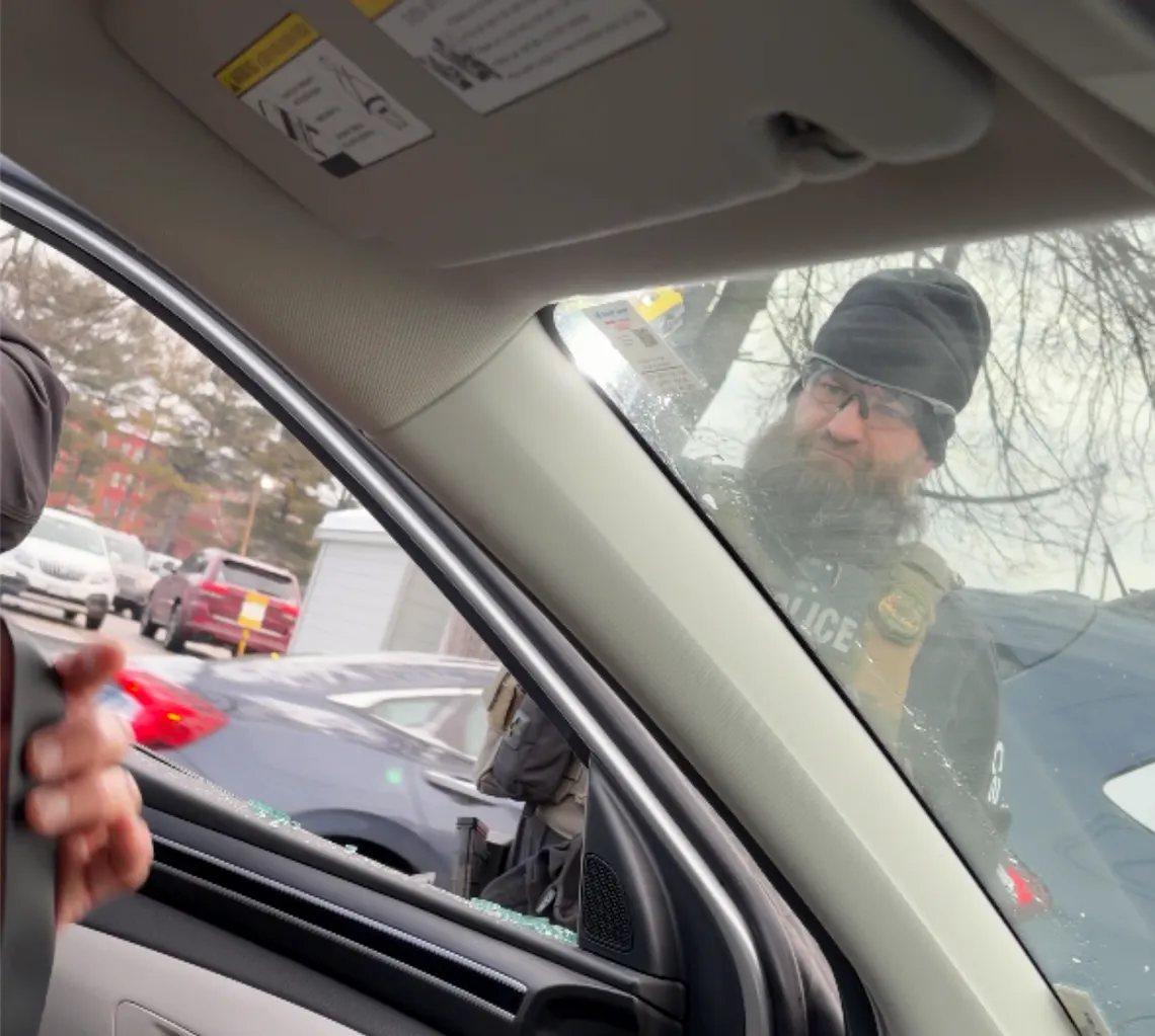 A photo shows a White man with glasses and a hat, wearing a uniform, standing in front of the driver's side of a car. A person's hand is visible inside the car. 