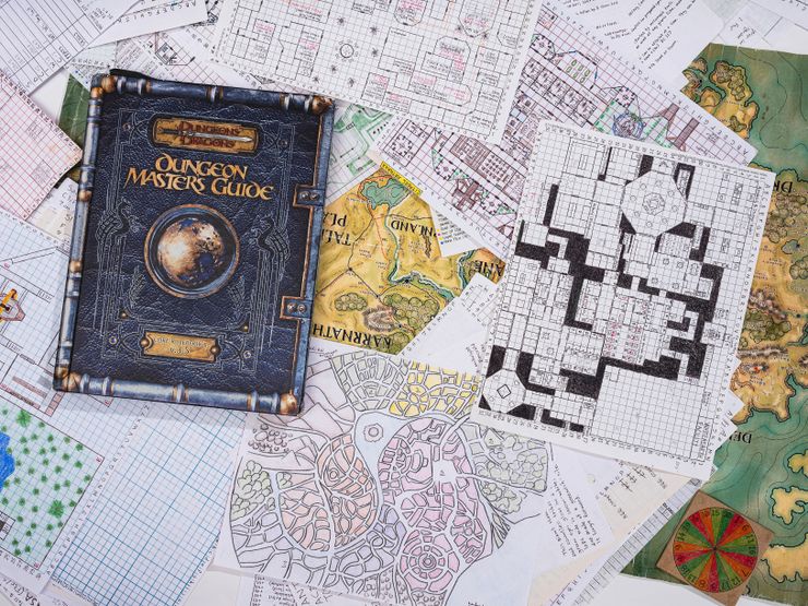 A Dungeons and Dragons book titled “Dungeon Master’s Guide” sits atop a pile of scattered maps and drawings. Many of the hand-drawn maps are sketched on graph paper. Some pages contain details in color, and others are drawn in black-and-white. 