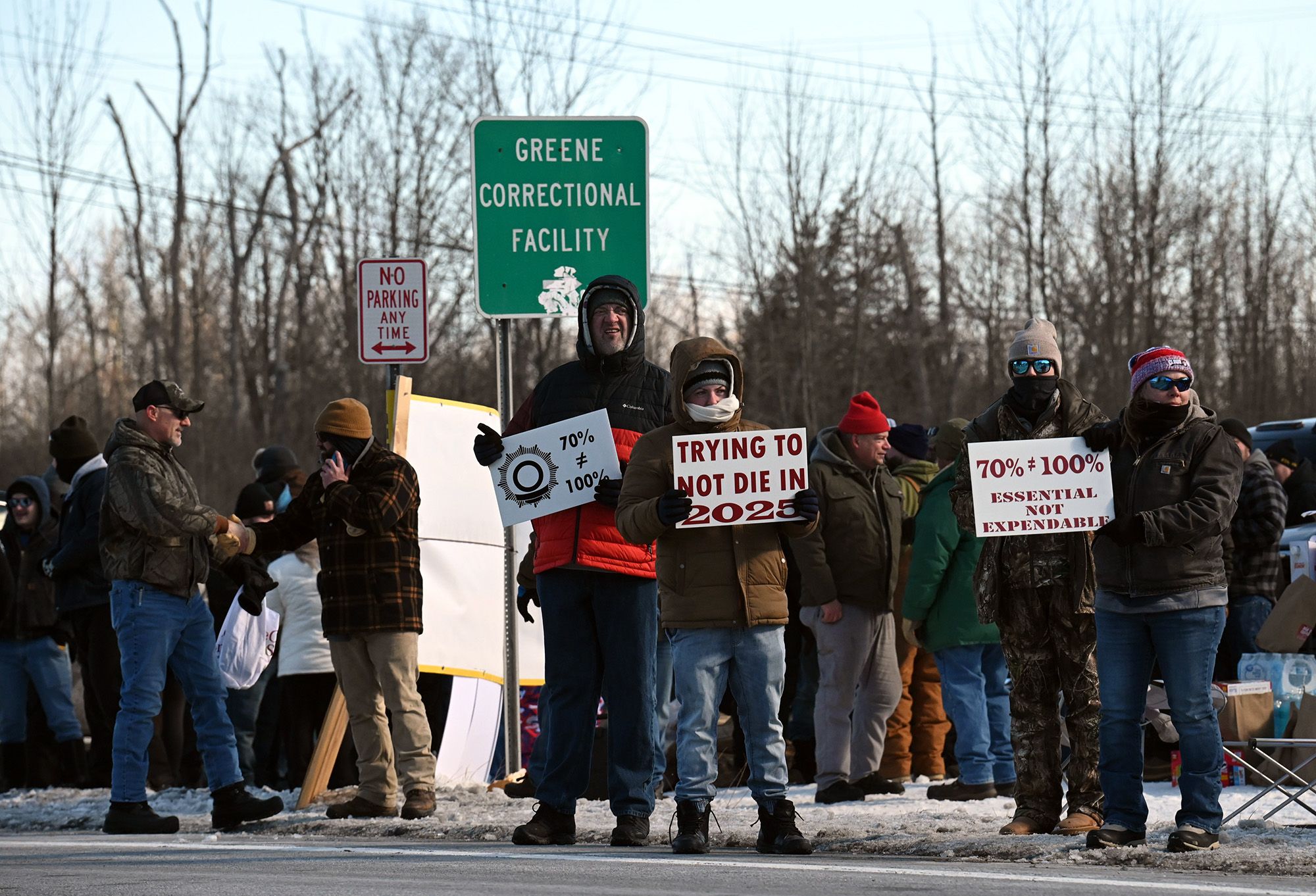 Protesters, wearing puffer jackets, beanies, gloves and pants, stand on the side of the road holding signs such as: "70% doesn't equal 100%," "Trying not to die in 2025," and "Essential not expendable." In the background, a road sign reads "Greene Correctional Facility." 