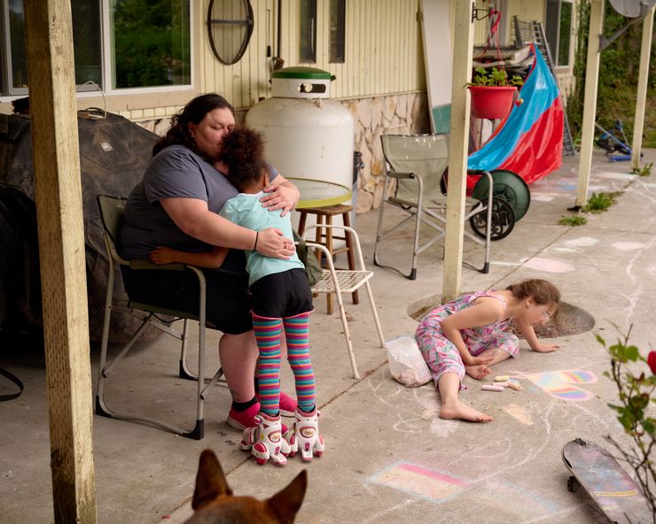 A woman with light-toned skin and a gray T-shirt is seated while hugging a young girl with a medium-toned skin as a young girl with light-toned skin plays on a patio.  