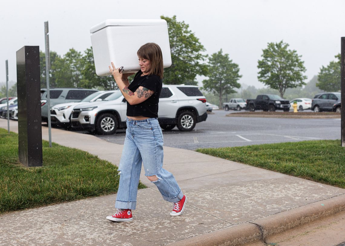 A photo of a White woman in a black t-shirt, blue jeans and red sneakers. The woman is carrying a white cooler while walking on a sidewalk on a rainy day. 