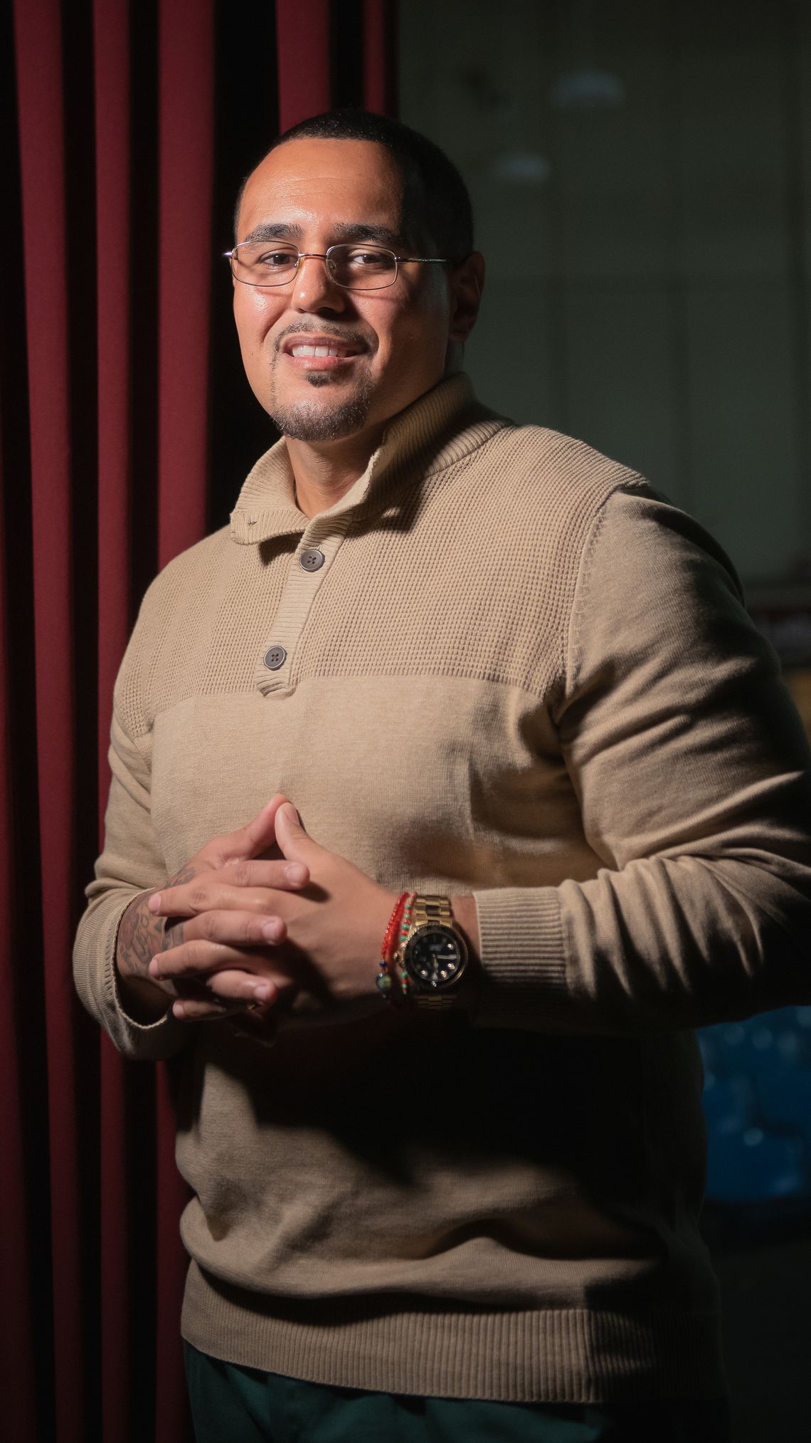 A Latino man with glasses and a thin goatee, wearing a tan sweater, stands with his hands interlocked at his waist, a slight smile on his face. 