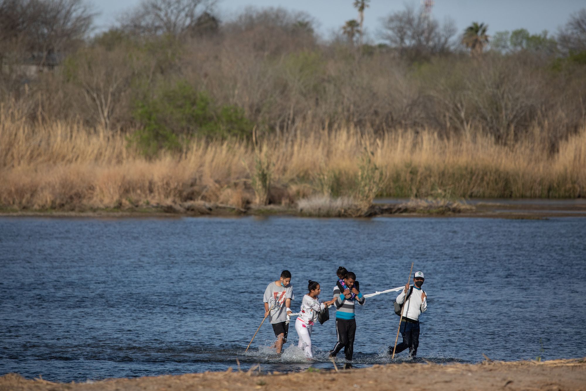 Three men and a woman wade across the Rio Grande River. The man second from the right has a small child on his shoulders. The man on the far right and the far left hold long sticks to help navigate the water.  