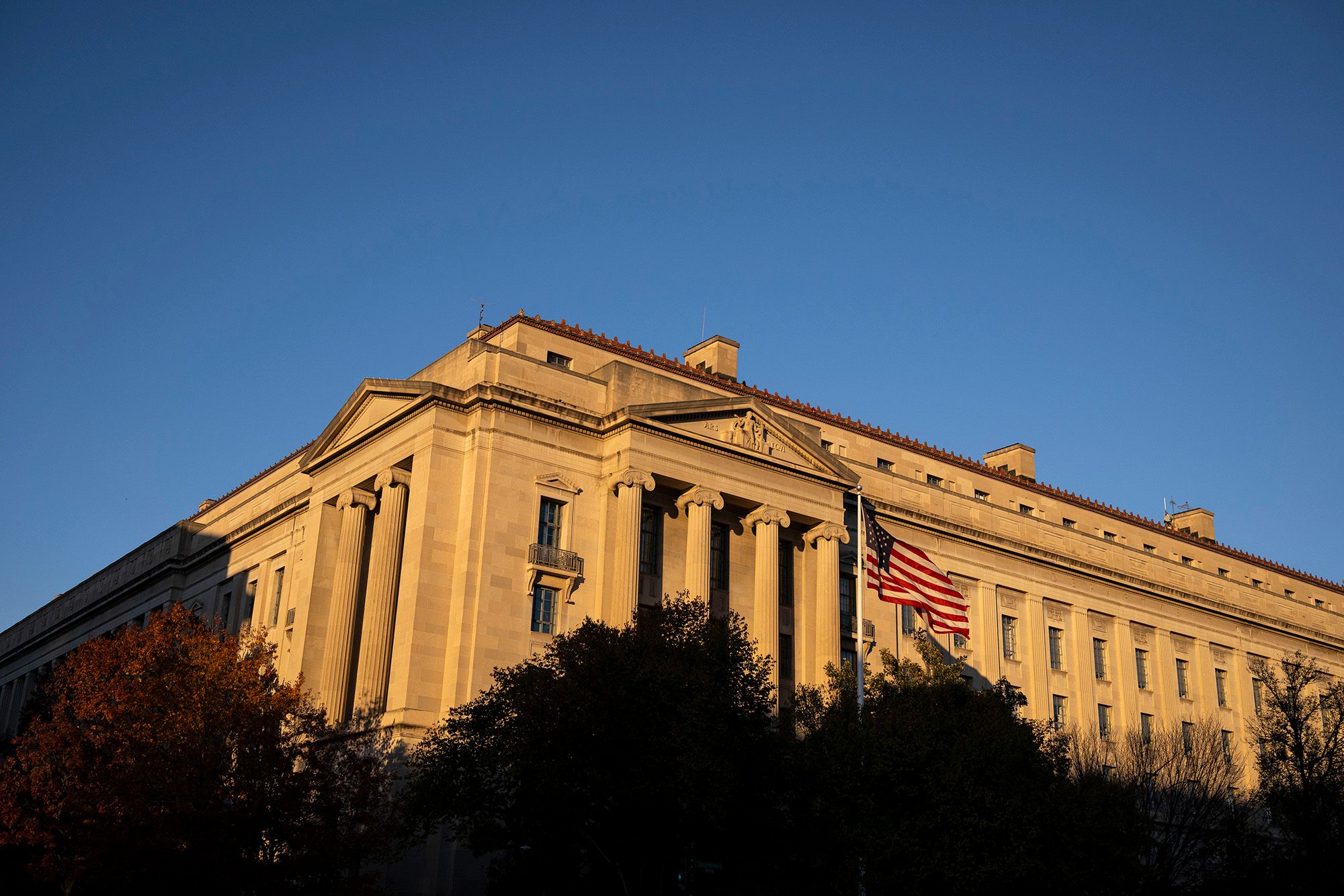 A color photo shows a gray stone building with columns. Dusk lighting makes the building appear yellow-orange. In the foreground are trees and an American flag on a pole.