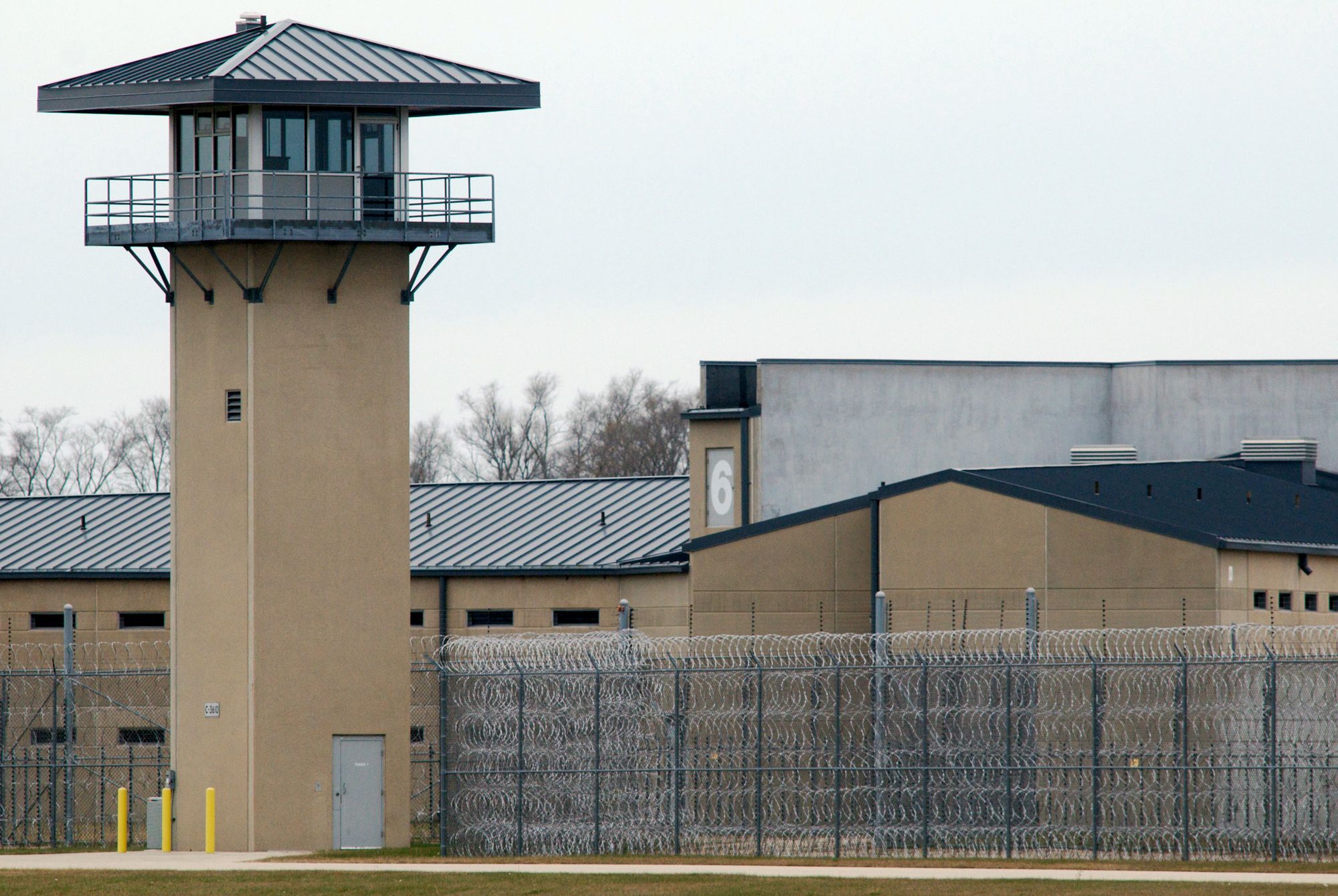 A tan guard tower and prison yard at an Illinois facility. There are wire fences and buildings in the background. 