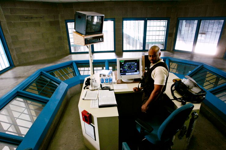 An officer stands in a control room with cells visible beyond the windows.  