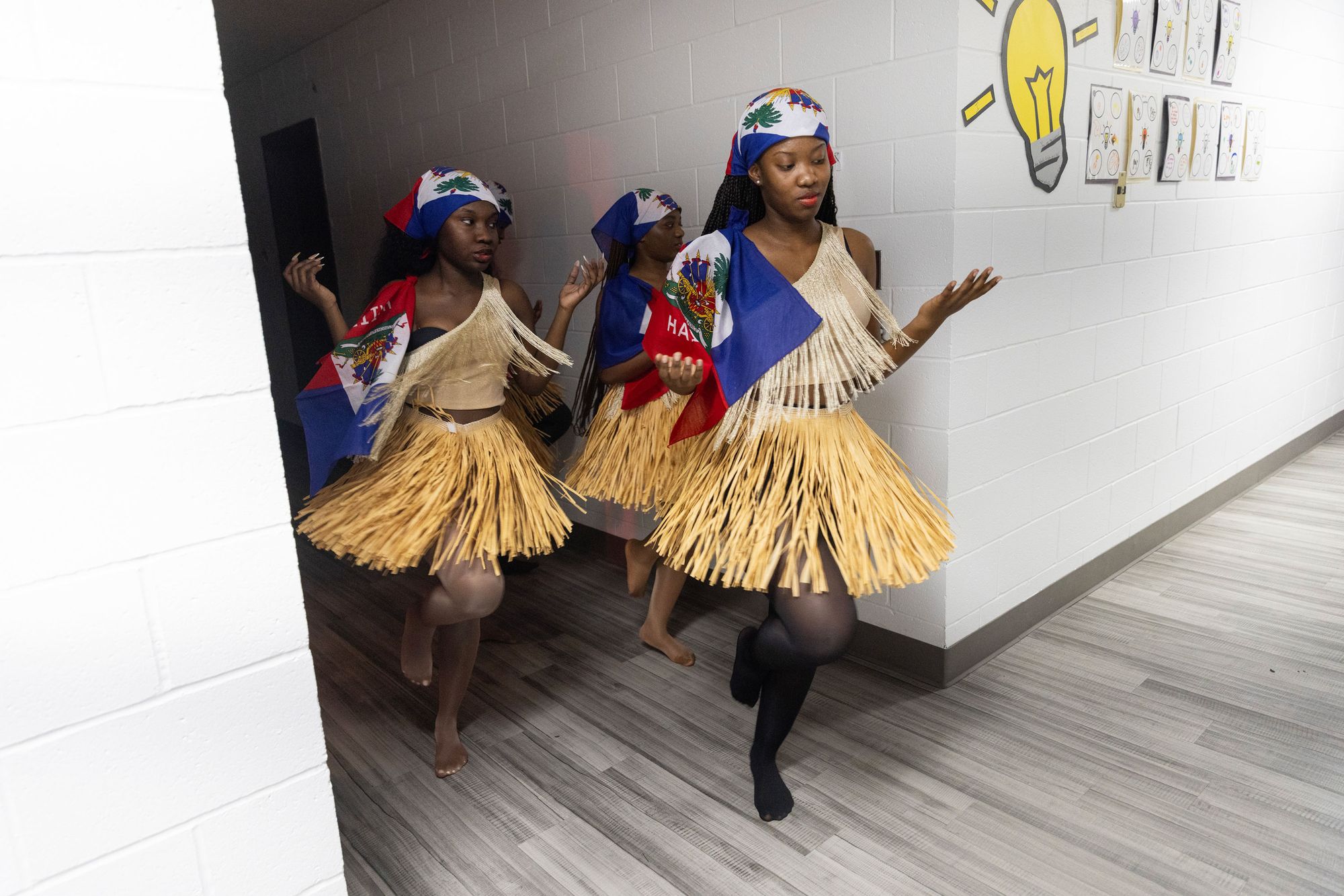 A photo shows a group of young Black women wearing grass skirts and tops and Haitian flags while dancing in a hallway. 