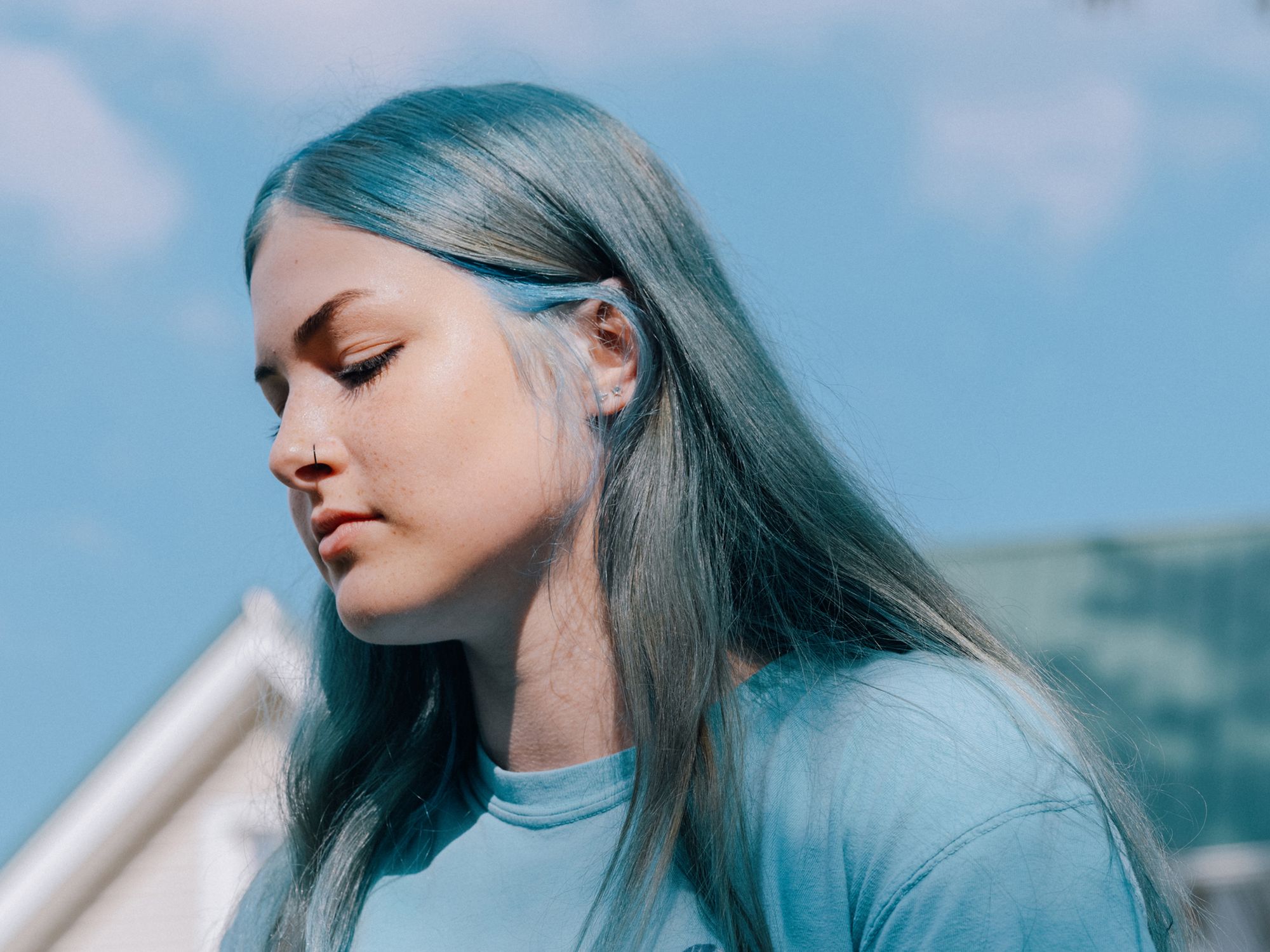 A photo shows a White woman with long blue hair, wearing a blue T-shirt, standing in front of a white building. Some leaves from a tree are visible above her. 