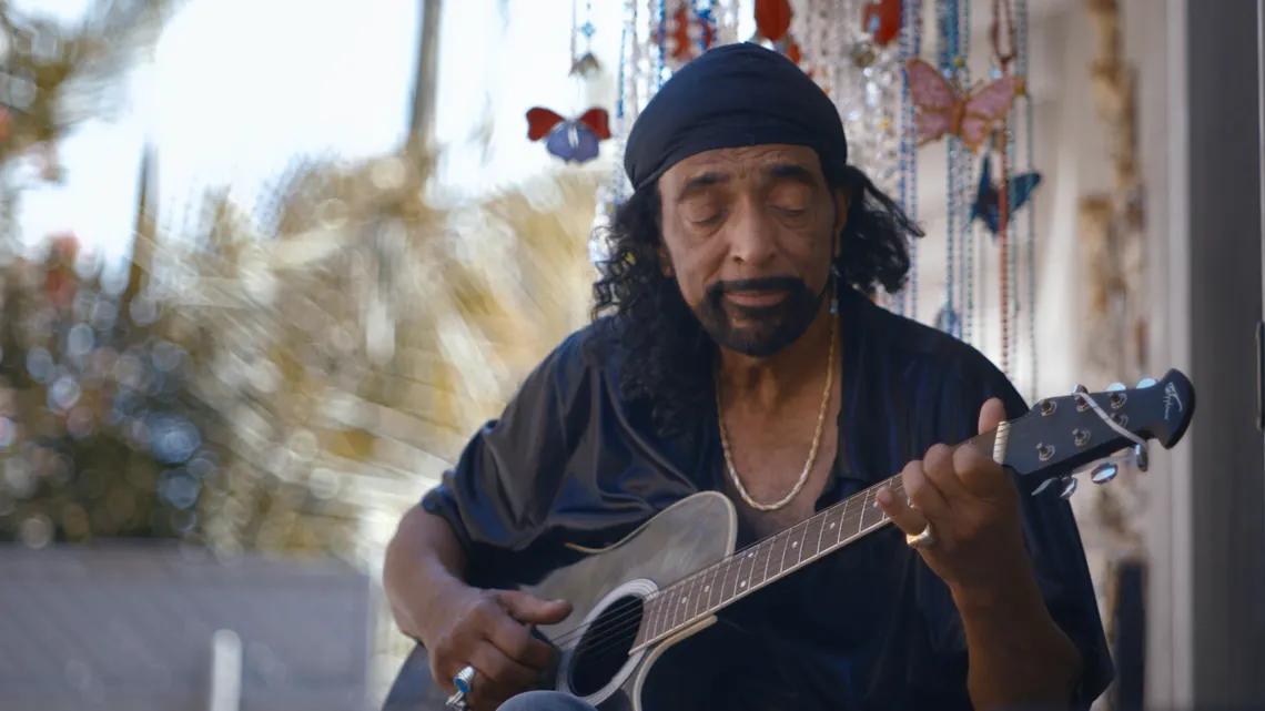 A photo shows Ike White, a Black man wearing a dark shirt. He is playing a guitar and sitting outside. 