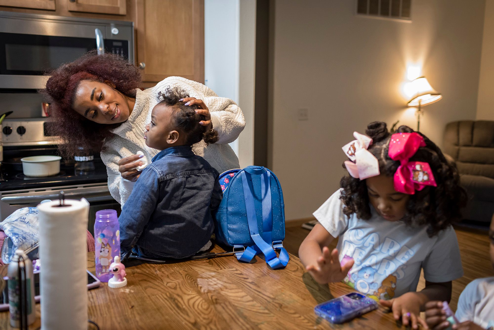 A photo shows a Black woman, with red hair and a white sweater, wiping the face of her young daughter while she sits on top of a kitchen table. Her eldest daughter, wearing pink hair ribbons and a blue Care Bears shirt, stands next to her while she looks at a phone placed on the table. 