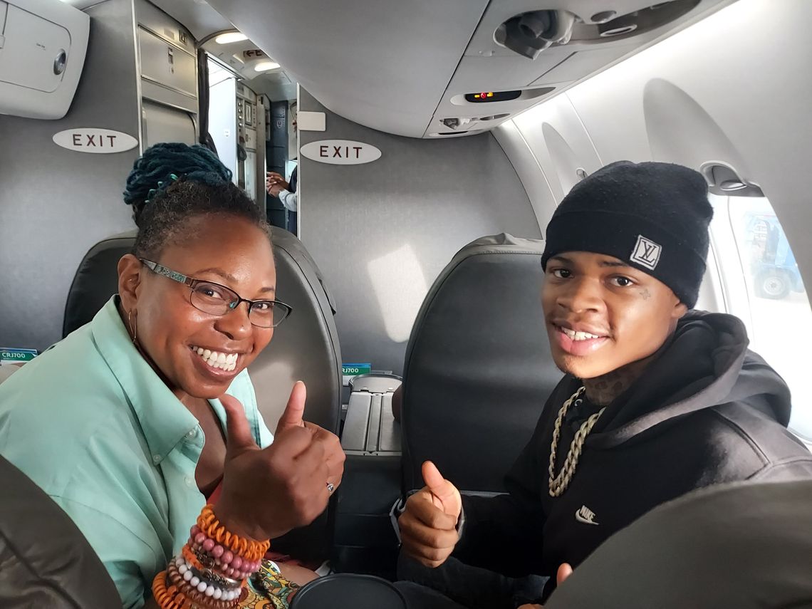A photo shows two people sitting in a plane: on the left, a Black woman with glasses and a pale green shirt, and on the right, a young Black man wearing a knit hat and a black sweatshirt. They are both smiling at the viewer with their thumbs up. 