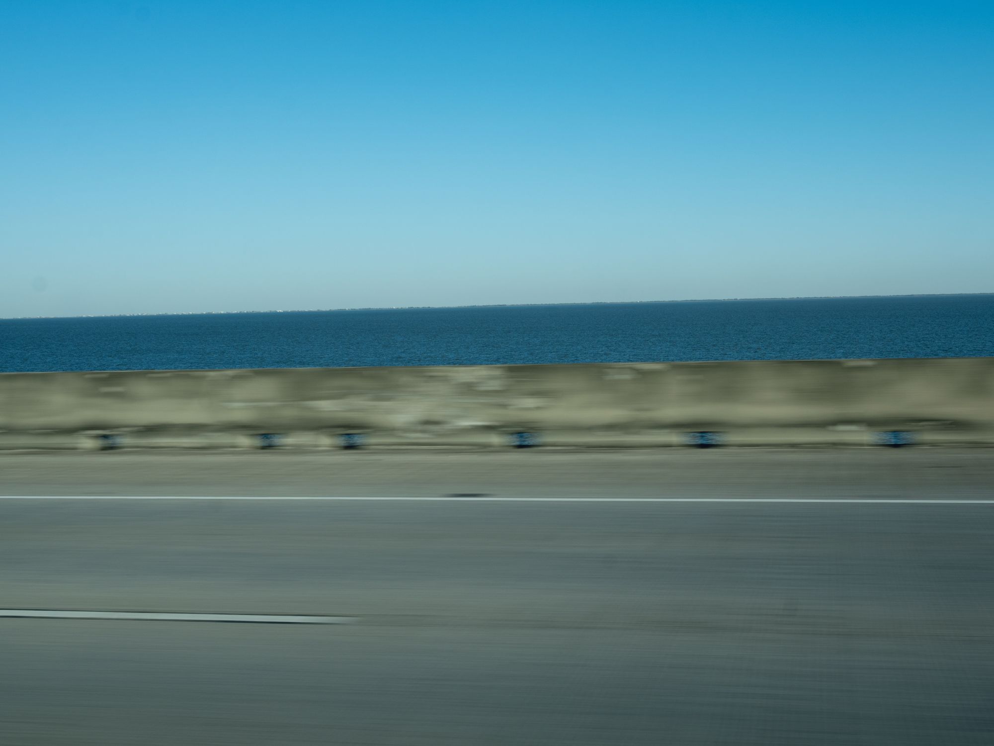 A blurred view of Lake Pontchartrain, the blue sky, and the road as seen from a moving vehicle.