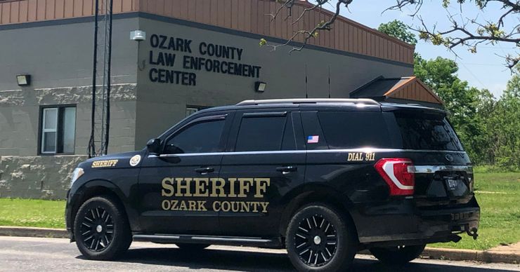 A Black SUV with the words “Sheriff Ozark County” on the driver’s door is parked in front of a gray brick building with a sign that reads “Ozark County Law Enforcement Center.” 