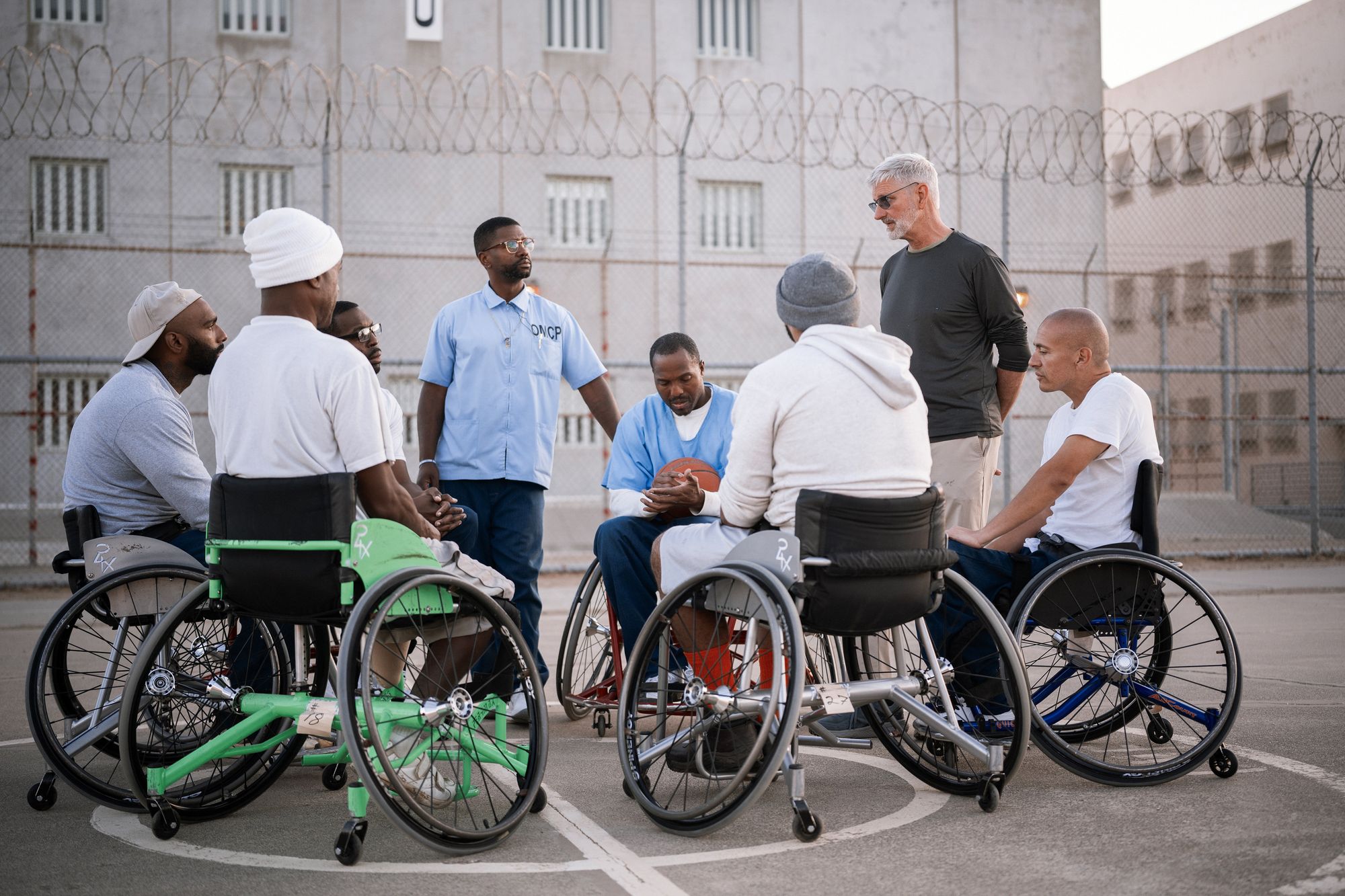 A photo shows six people gathered, either standing or in wheelchairs, around Coach Jeremiah Holland, a White man. The people have different skin tones ranging from dark-toned to light-toned. 