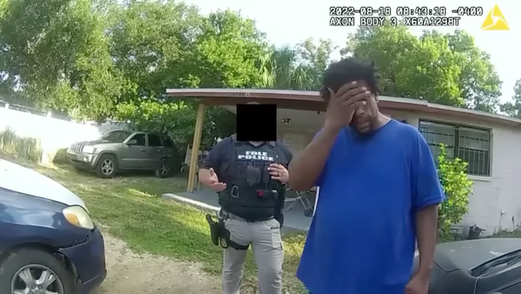 A Black man in a blue T-shirt covers his eyes with one hand as he stands in his front yard. A police officer, wearing protective gear with the words "FDLE Police" on it, stands left of him in the background. 