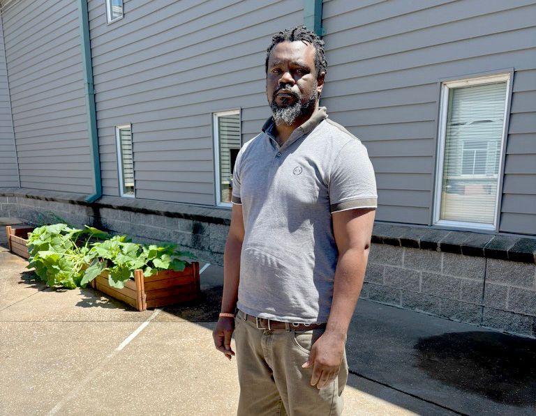 A photo shows a Black man with a beard standing in front of a gray building, posing for a portrait. He wears a light-colored top, khaki pants and blue sneakers.