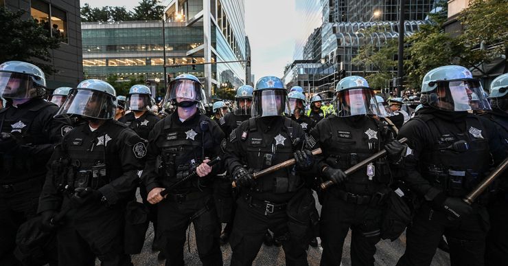 A photograph shows a crowd of police officers, wearing riot gear and holding batons, standing in formation in the middle of a street. 