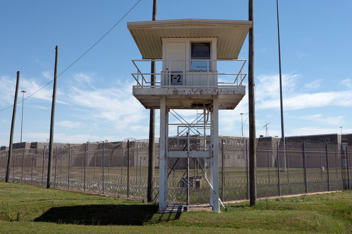A photo shows a guard tower in front of an area and low buildings surrounded by barbed wire fencing.