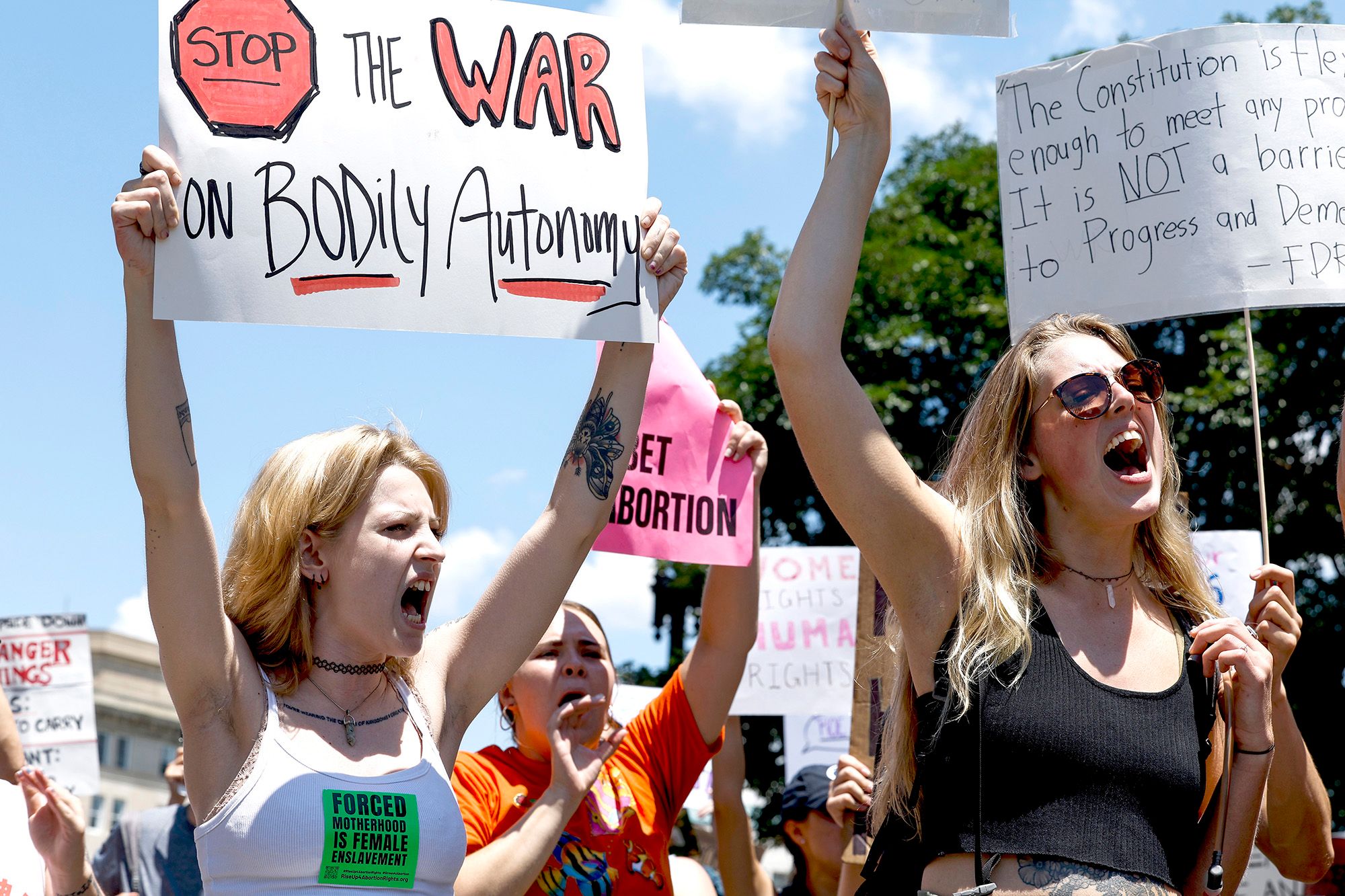 A White woman, wearing a white tank top and a label that reads "Forced motherhood is female enslavement," yells while holding a sign that reads, "Stop the war on bodily autonomy." People are protesting in the background. 