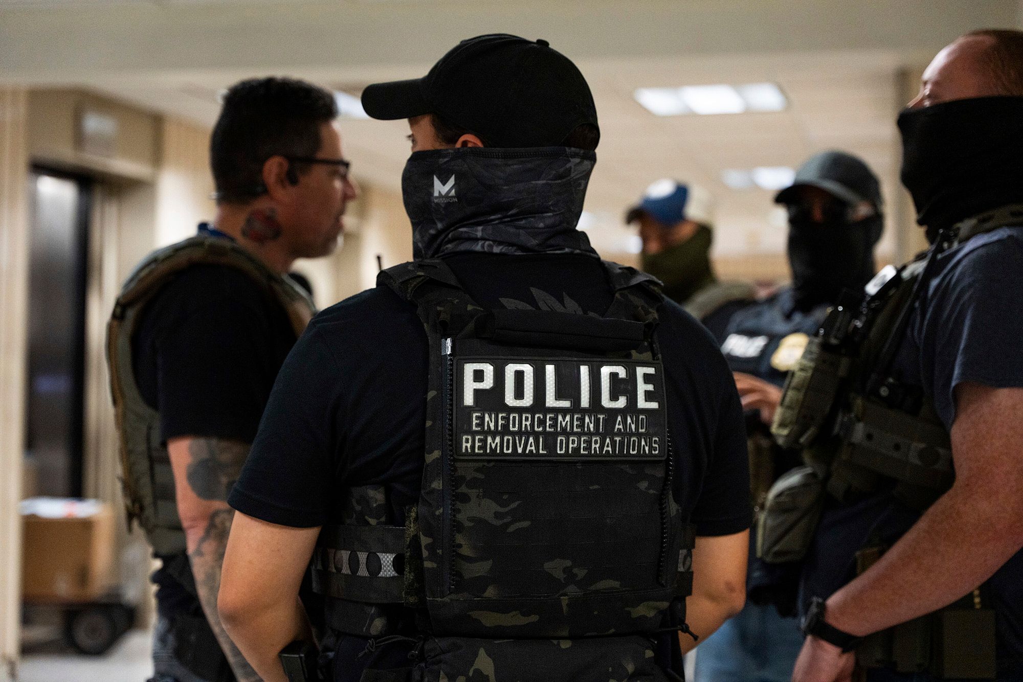 A photo shows the back of a light-skinned police officer wearing a black T-shirt, a black cap, face covering and a vest with the words "police enforcement and removal operations." There are 4 officers standing in the background.  