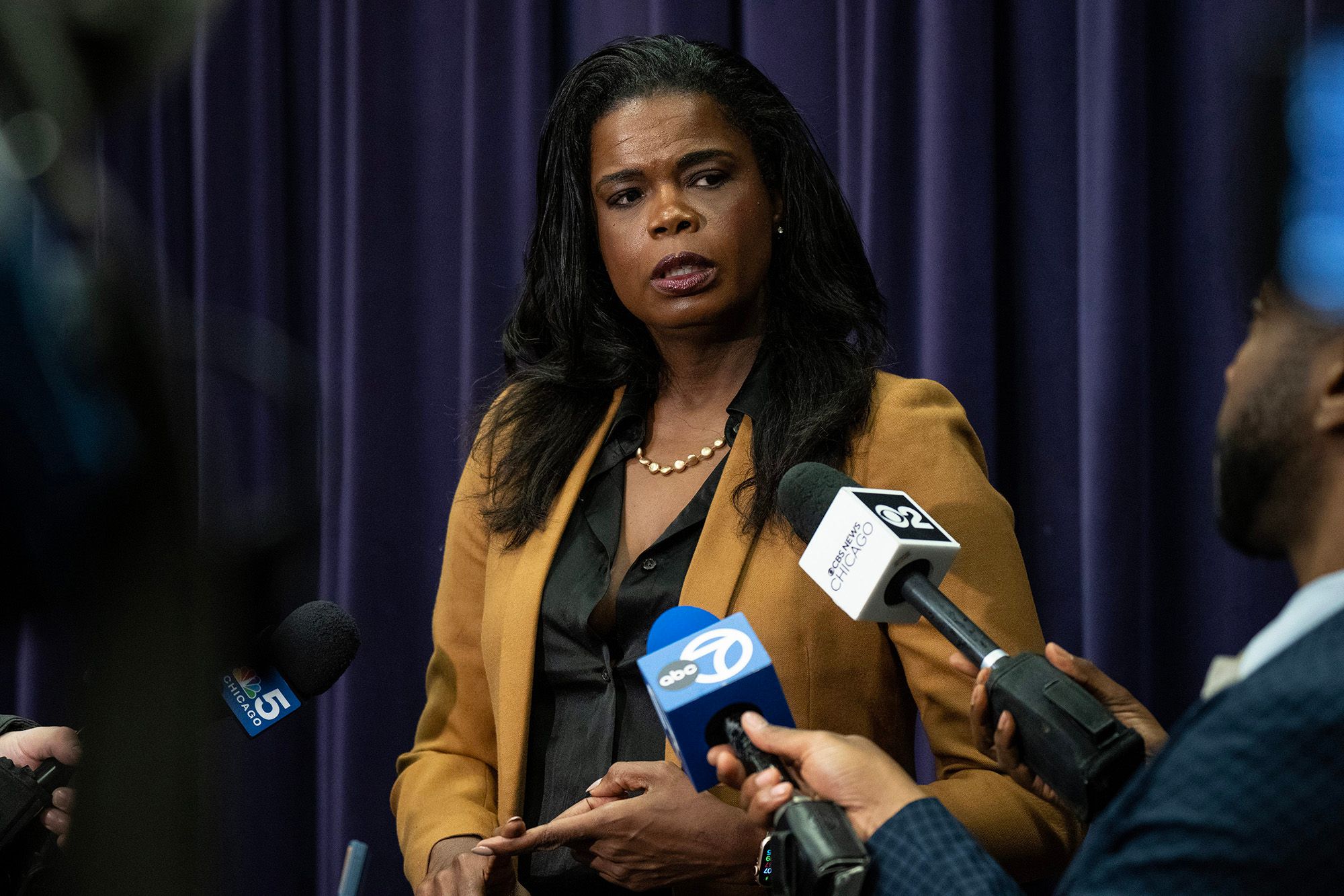 Kim Foxx, a Black woman wearing a tan suit, speaks into mics held by reporters surrounding her. 