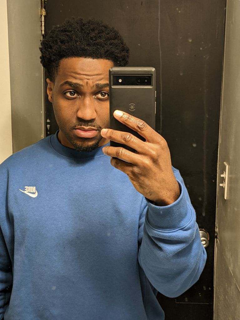 Darrian Bennett, a Black man with short hair and a blue long-sleeved sweatshirt, takes a selfie. 
