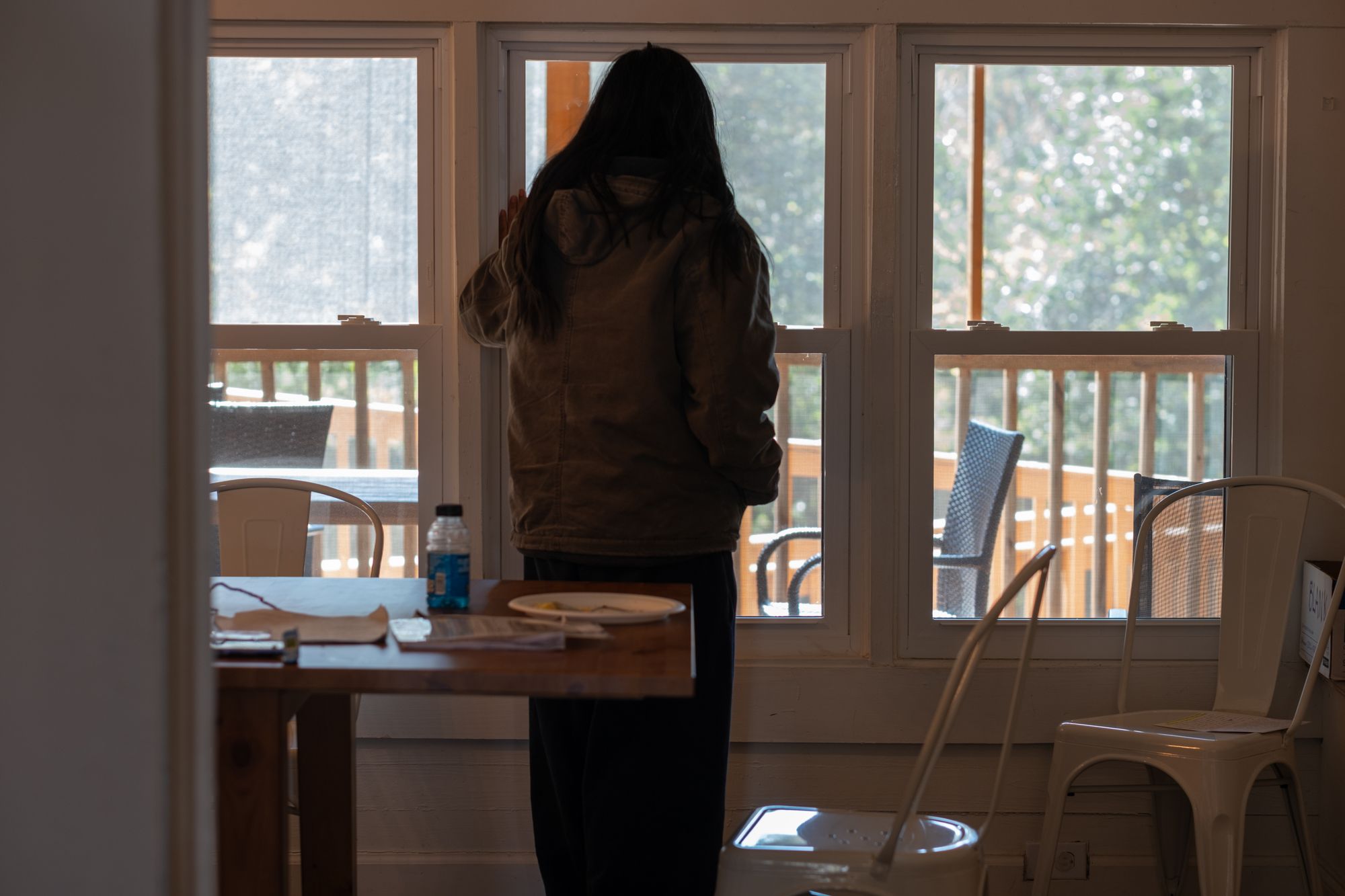 A woman with long hair faces away from the camera, silhouetted in front of three windows.