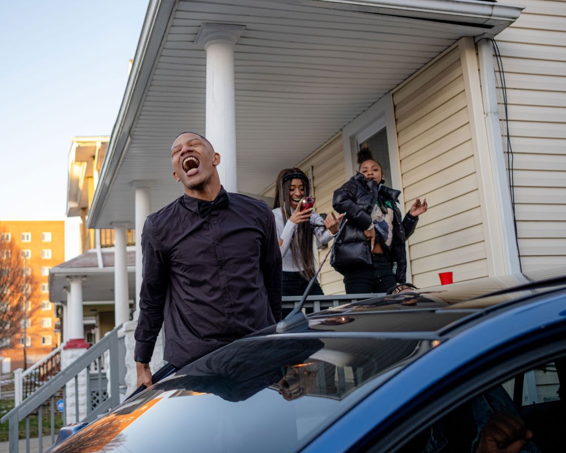 A photo of a young Black man singing next to a car while two young women behind him dance and record from the porch of a house. 