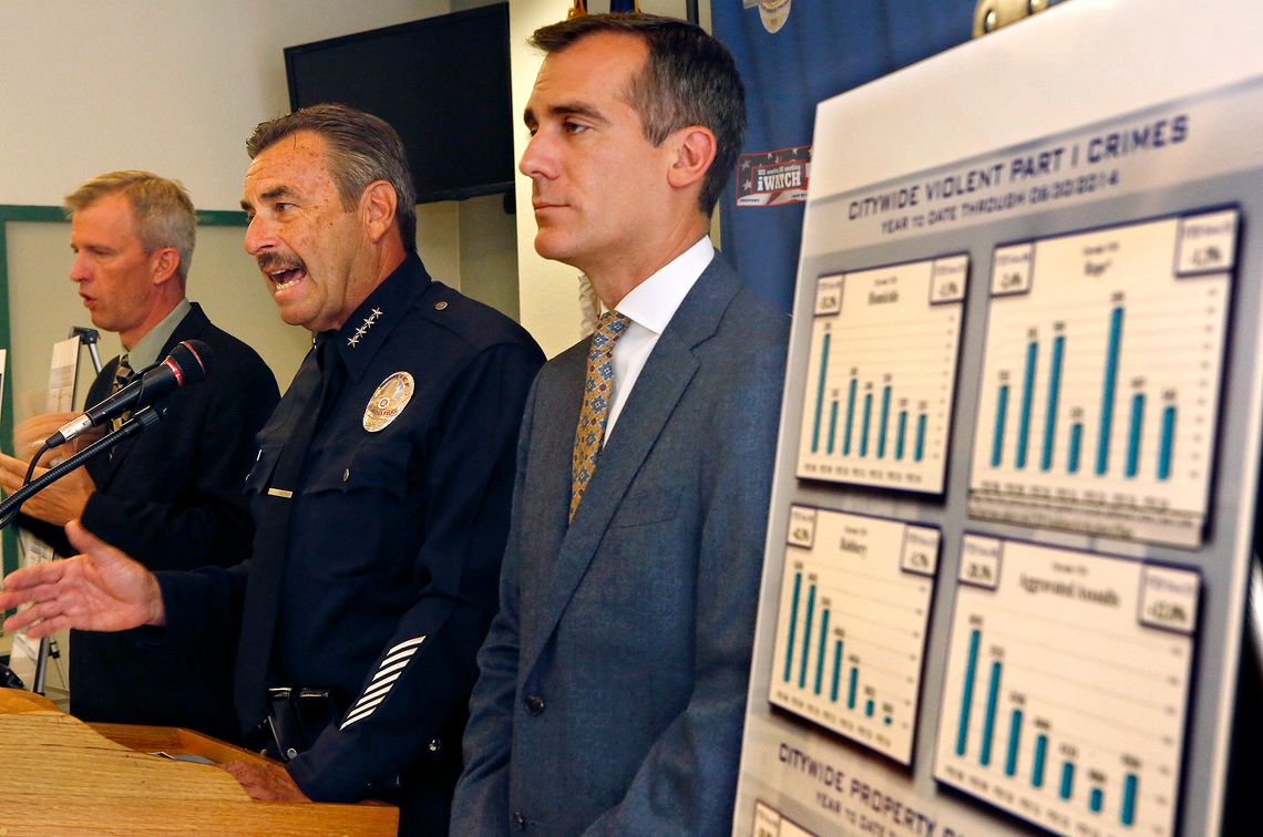A photo shows a White man in a police uniform speaking into a microphone at a podium. There are White men in suits on either side of him. On the right side of the photo, a poster board displays bar graphs related to crime data.  