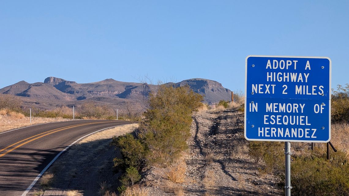 A photo shows a blue highway sign that reads, “Adopt A Highway, next 2 miles. In Memory of Esequiel Hernandez.” There was a landscape with mountains and a 2-lane road in the background. 