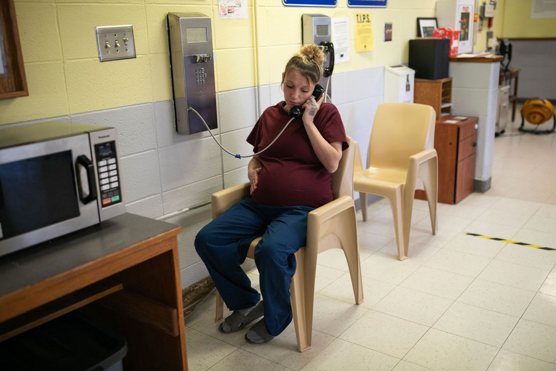 A photo shows a pregnant White woman with blond hair wearing a prison uniform, sitting and talking on the phone.