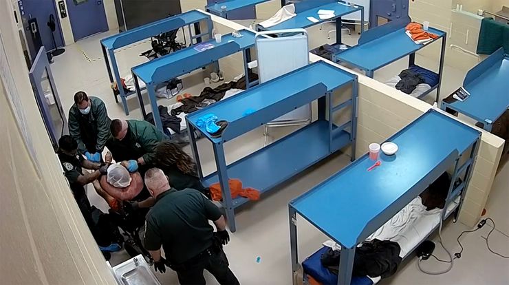 Five jail staff surround a man restrained in a wheelchair, with his head covered in a mesh hood.