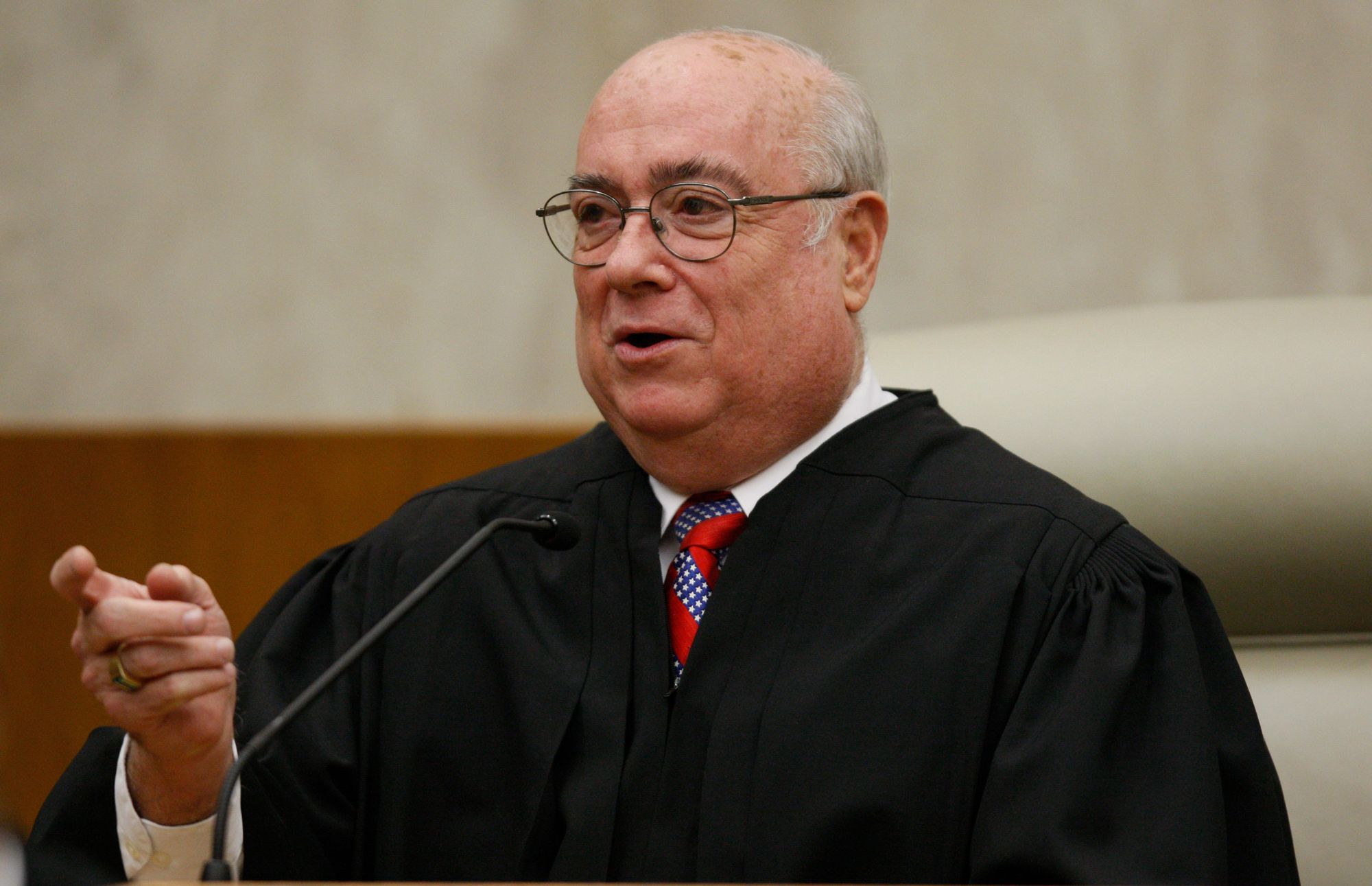 A photo of a White man behind a microphone gesturing while wearing a black judge’s robe. 